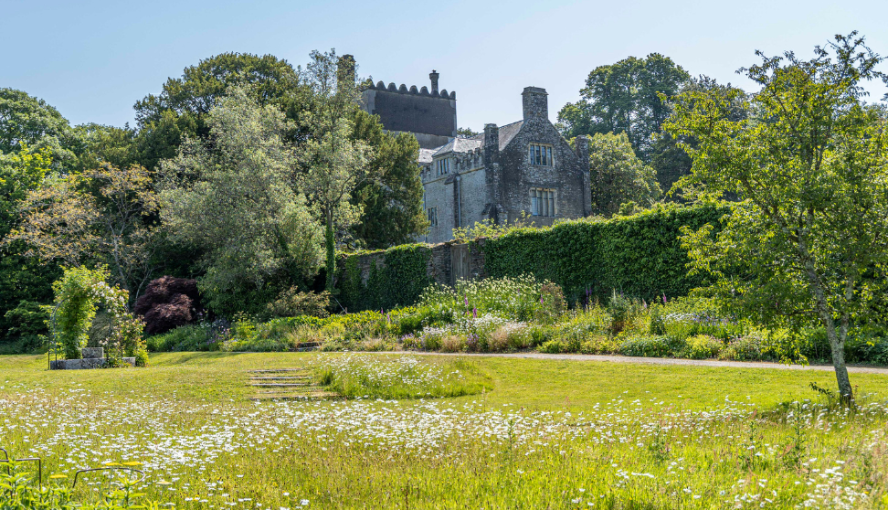 Historic stone house overlooking a wildflower meadow and formal gardens on a sunny day.
